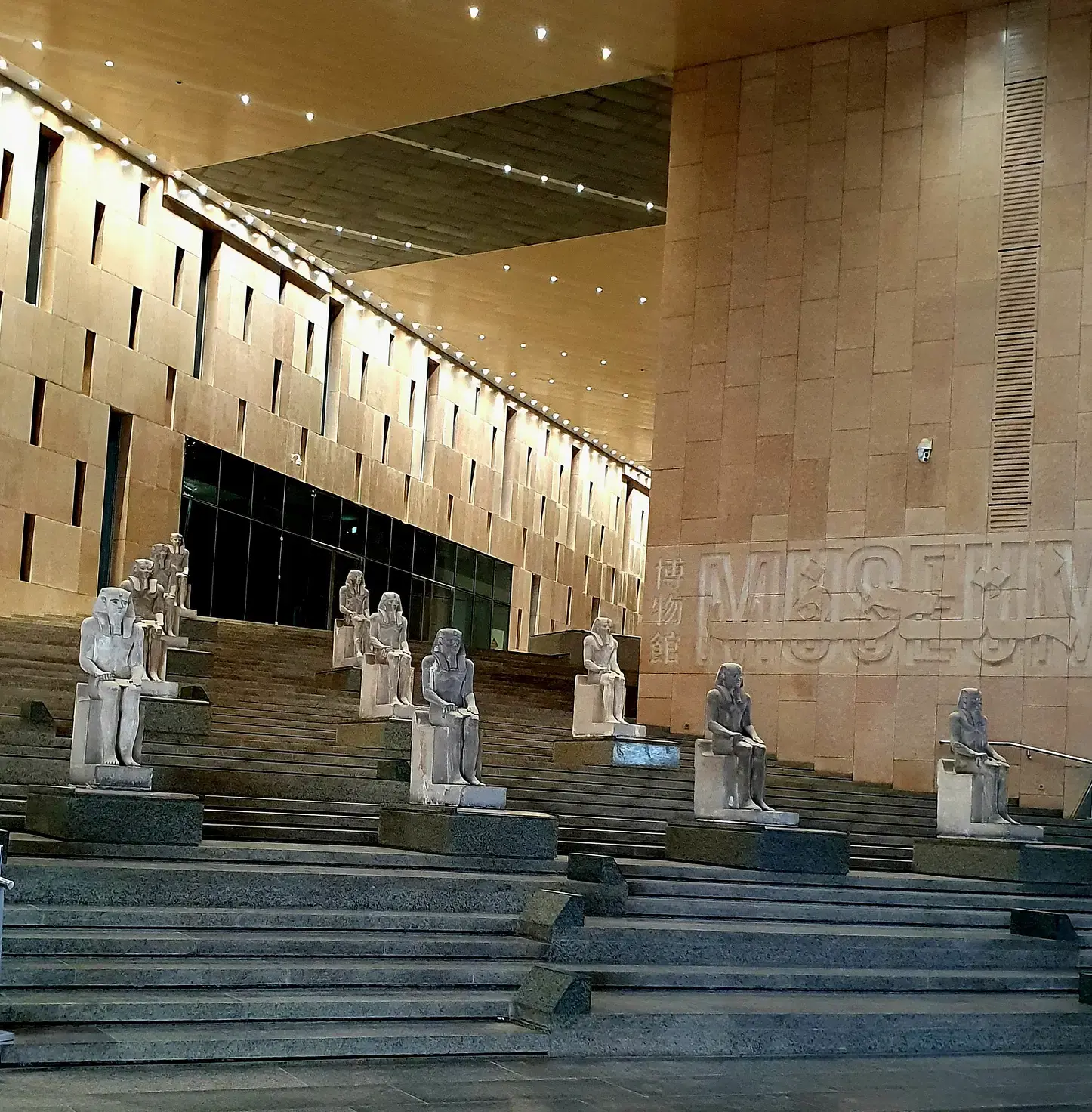 Visitors viewing ancient Egyptian artifacts, including statues and mummies, at the Grand Egyptian Museum in Giza, with the Great Pyramids in the background.