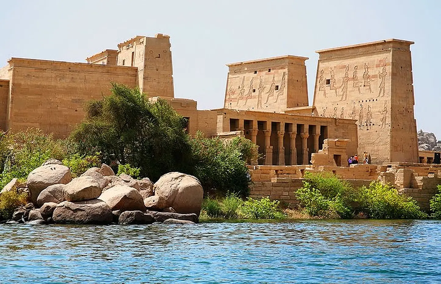 A traditional felucca sailboat gliding across the serene waters of the Nile in Aswan, with lush greenery and historical sites in the background.