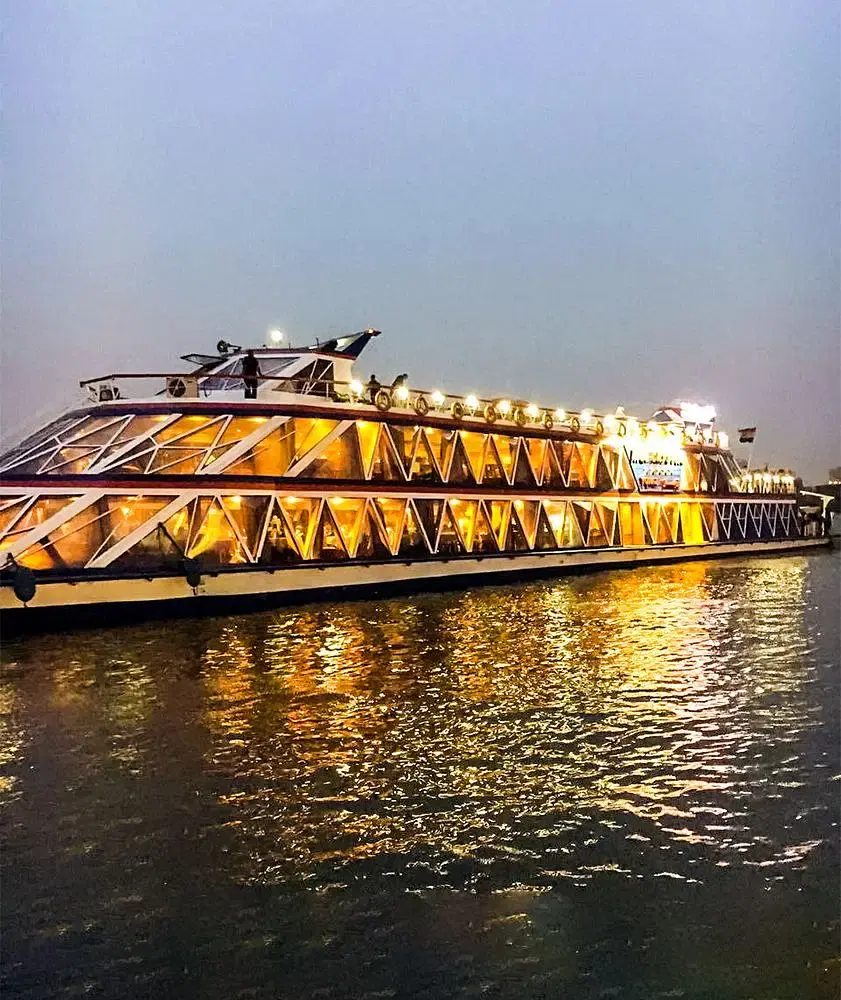 A nighttime view of the Nile River with a dinner cruise boat illuminated with lights, passengers enjoying a walk along the riverside in Cairo, and the city skyline in the background.