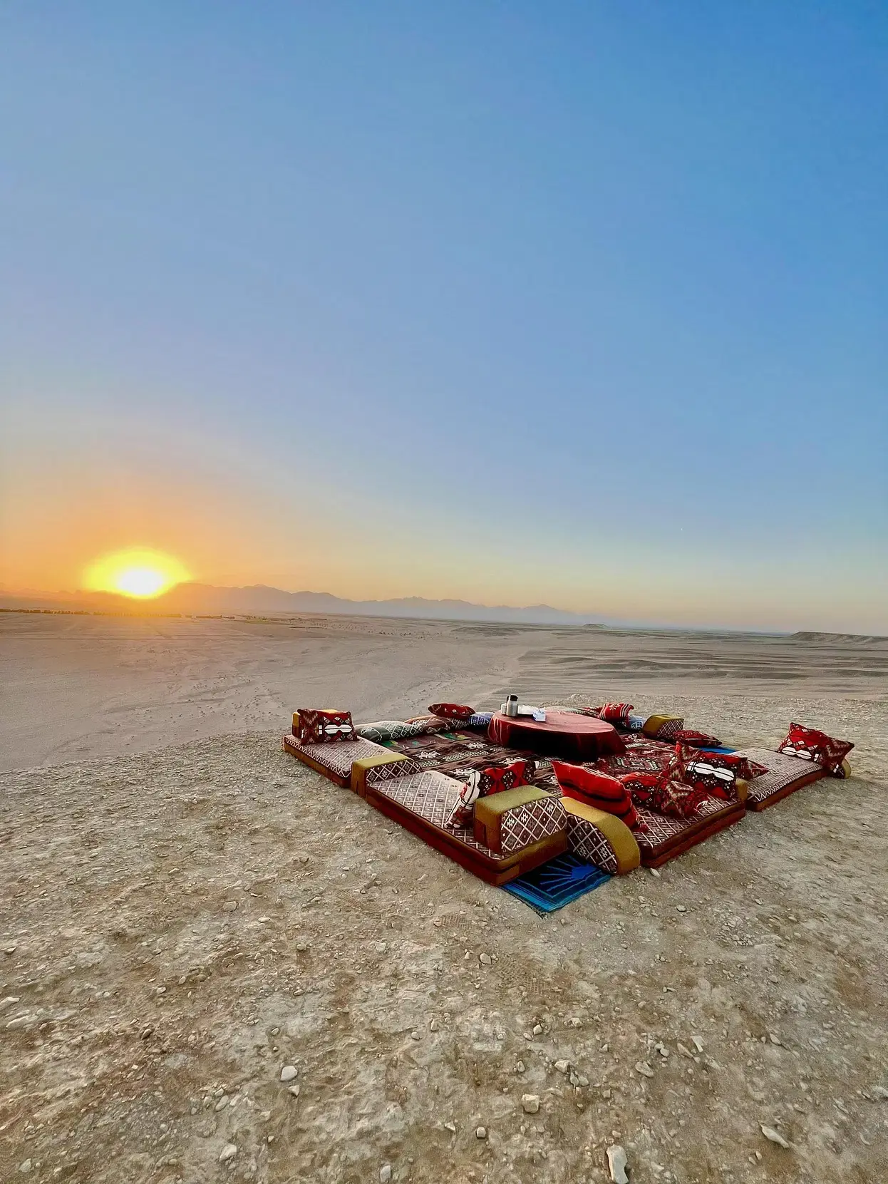 Tourists riding camels and 4x4 vehicles across the vast desert landscape near Hurghada, with golden sand dunes and a clear blue sky in the background.